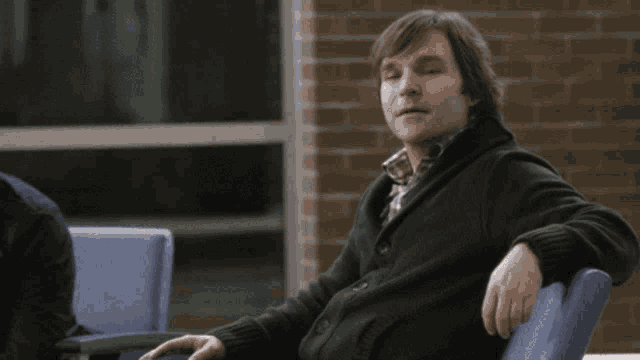 a man in a black sweater sits in a blue chair in front of a brick wall