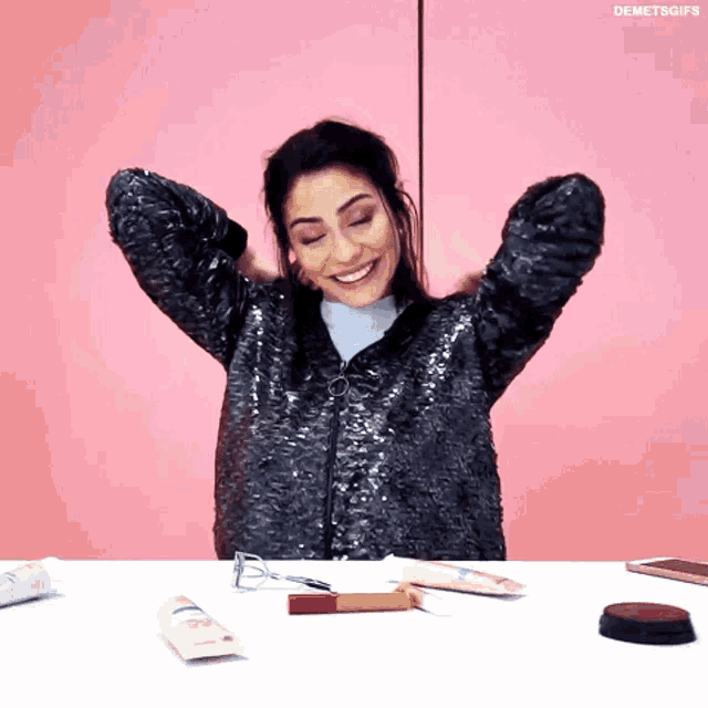 a woman in a sequined jacket is sitting at a table with her arms outstretched and smiling .