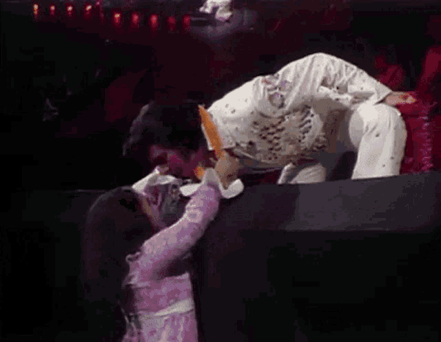 elvis presley is kneeling down on stage while a little girl reaches out to him .