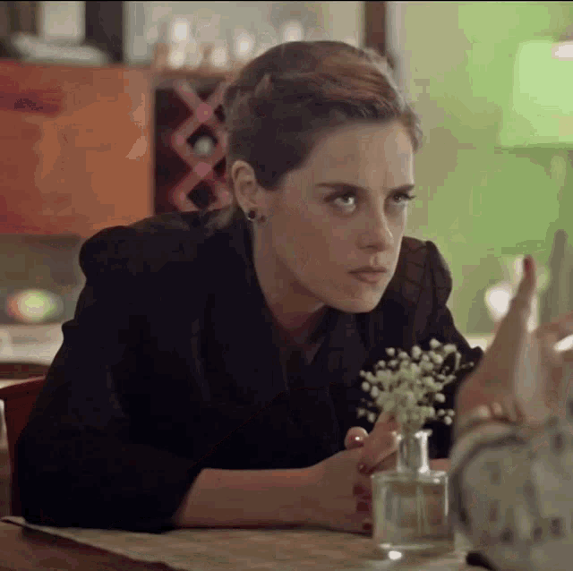 a woman in a black jacket sits at a table with a vase of baby 's breath
