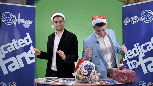 two men wearing santa hats are standing in front of a sign that says " cated fans "