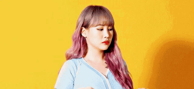 a woman with purple hair and a blue shirt is standing in front of a yellow wall .