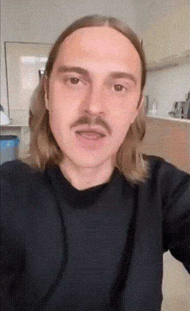 a man with long hair and a mustache is taking a selfie in a kitchen .