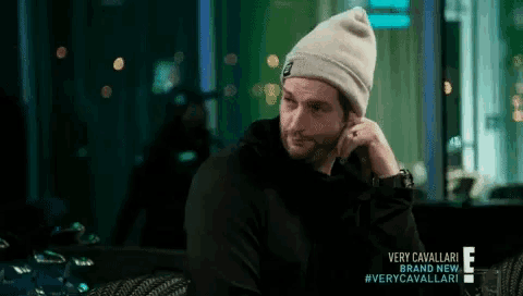 a man wearing a beanie is sitting in a dark room with a very cavallari brand new advertisement behind him