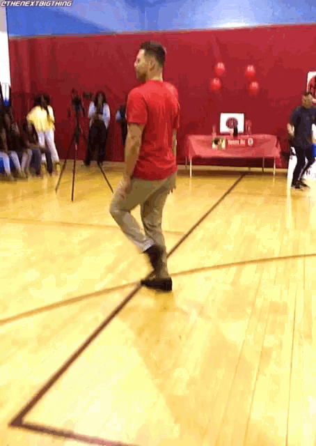 a man in a red shirt is dancing on a basketball court in front of a red wall