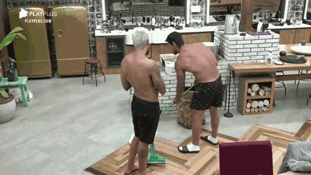 two men without shirts are cleaning a wooden floor with a mop in a kitchen with playplus.com in the corner