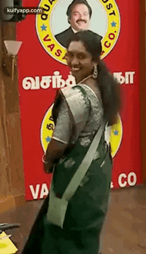 a woman in a green saree is standing in front of a red sign with a picture of a man on it .