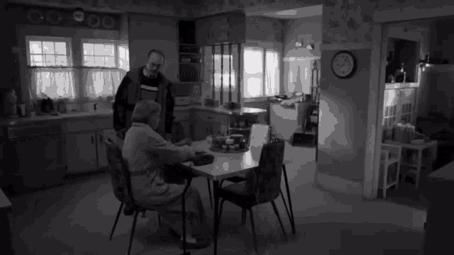 a black and white photo of two people in a kitchen with a clock on the wall