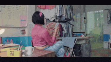 a woman in a pink shirt sits at a desk looking at a phone