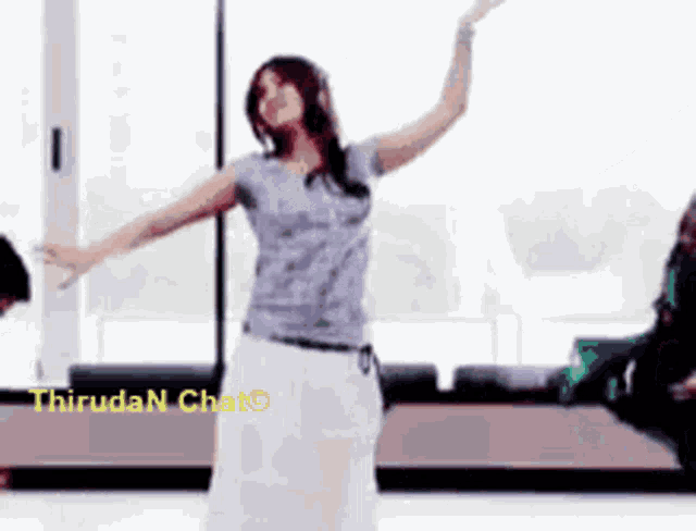a woman in a gray shirt and white skirt is dancing in front of a window with the words thiruda n chat visible