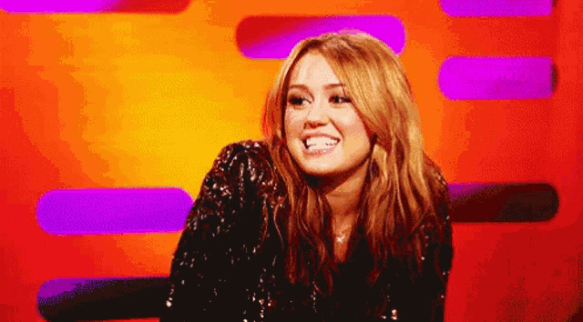 a woman in a sequined jacket smiles in front of a purple and orange background