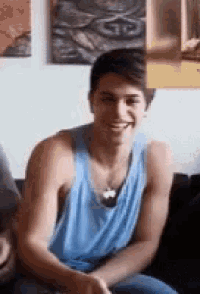 a young man is sitting on a couch wearing a blue tank top .