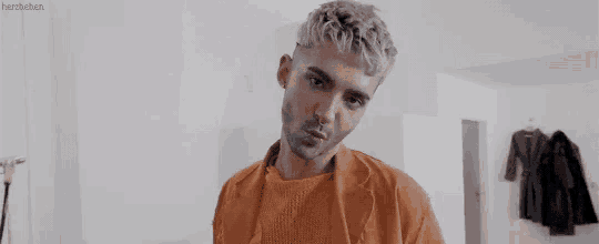 a man with gray hair and a beard is standing in a room wearing an orange jacket .