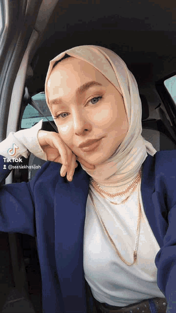 a woman wearing a hijab and a blue jacket is sitting in a car with tiktok written on the bottom right