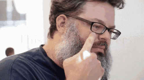 a man with glasses and a beard is pointing his finger at his nose