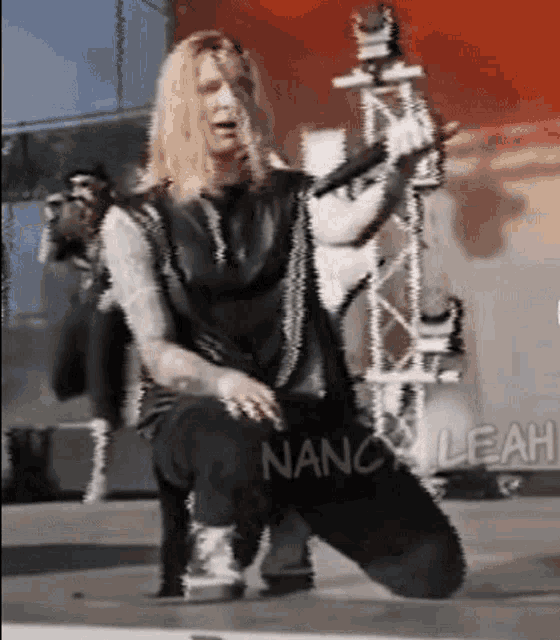 a man with long blonde hair is kneeling down and singing into a microphone with nancy leah written on the bottom right