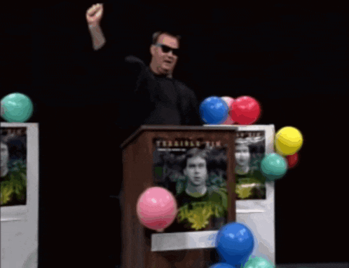 a man stands at a podium surrounded by balloons and a poster that says " terrible tik "