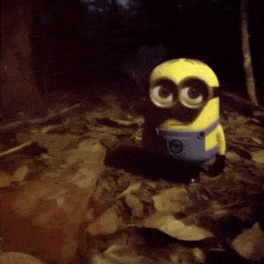 a toy minion is standing in a pile of leaves in the woods .