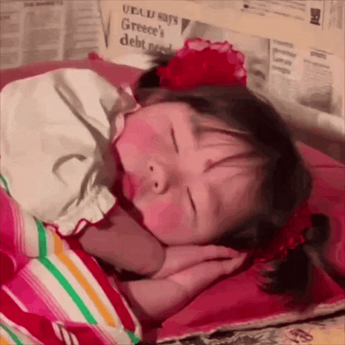 a little girl sleeping next to a newspaper that says greece 's debt crisis