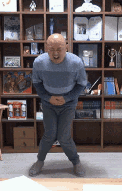 a bald man in a blue sweater is standing in front of a bookshelf holding his belly