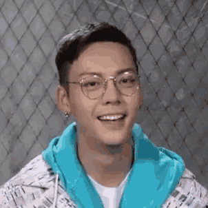 a young man wearing glasses and a blue jacket is smiling while standing in front of a chain link fence .