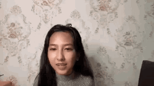 a woman is sitting in front of a wall with a floral wallpaper and looking at the camera .