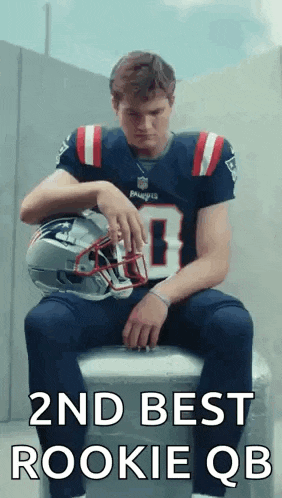 a man in a patriots jersey is sitting on a cooler holding his helmet .