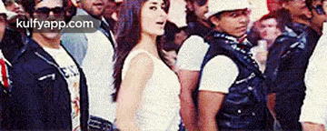 a woman in a white tank top is standing in front of a crowd of people with the website kulfyapp.com visible