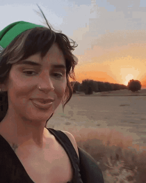 a woman wearing a green hat and a black tank top smiles in front of a sunset