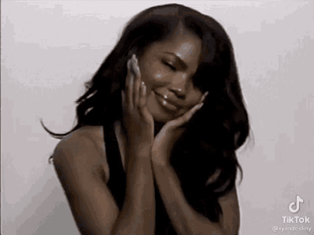 a woman with long black hair is smiling and holding her face with her hands .