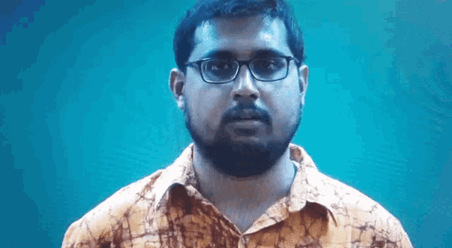 a man with glasses and a beard looks at the camera with a blue background