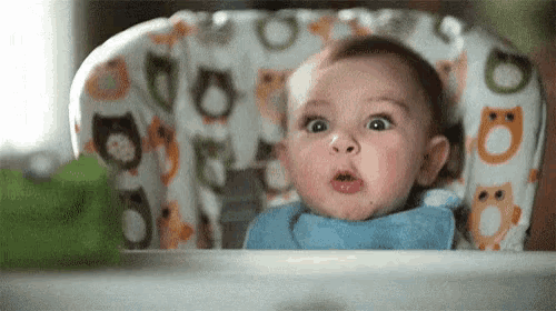 a baby is sitting in a high chair with a bib on and making a funny face .