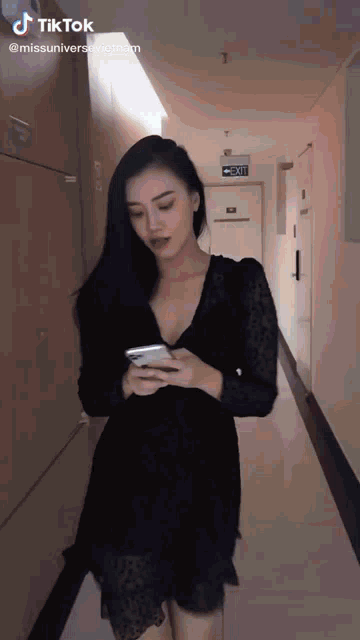 a woman in a black dress is walking down a hallway looking at her phone