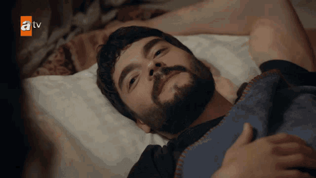 a man with a beard is laying on a bed with a tv logo on the bottom