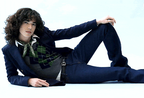 a young man with curly hair is laying on the floor wearing a blue jacket and jeans