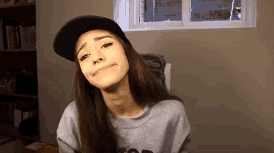 a girl wearing a baseball cap and a grey sweatshirt is making a funny face .