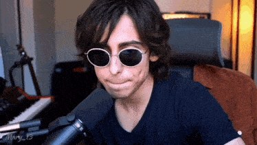 a man wearing sunglasses and a black shirt is sitting in front of a microphone .