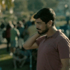a man with a beard is standing in a park with his hand behind his head