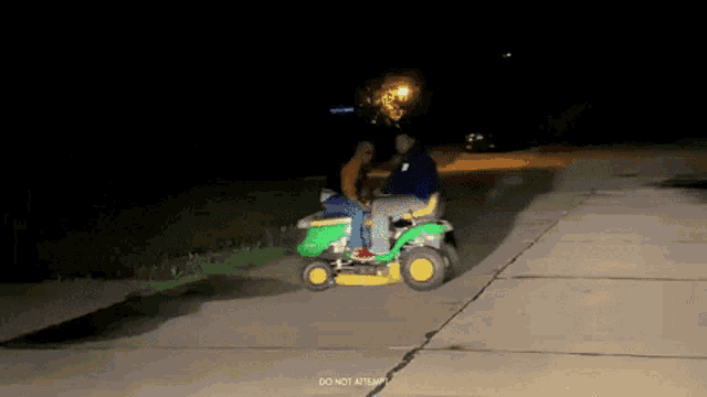 two men are riding a john deere lawn mower on a sidewalk at night