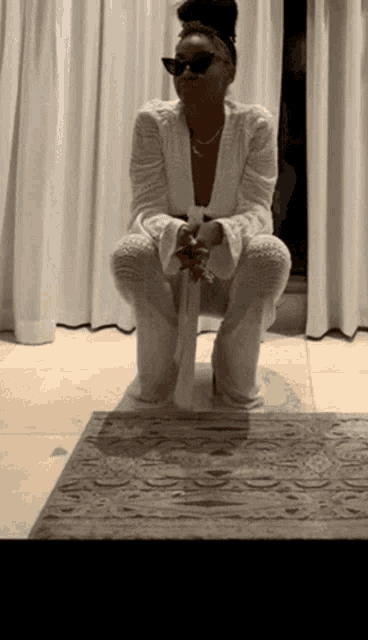 a woman wearing sunglasses is squatting down in front of white curtains