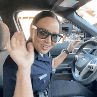 a woman in a police uniform is waving from the driver 's seat of a car .
