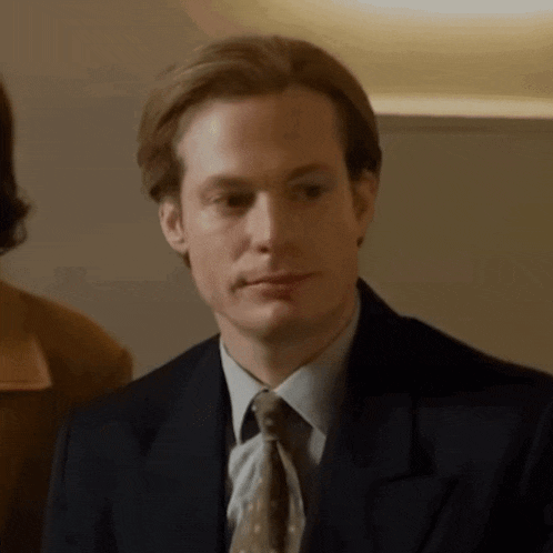 a man in a suit and tie is looking at the camera with a woman in the background .