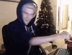 a man in a hoodie is sitting in front of a christmas tree with his arms outstretched