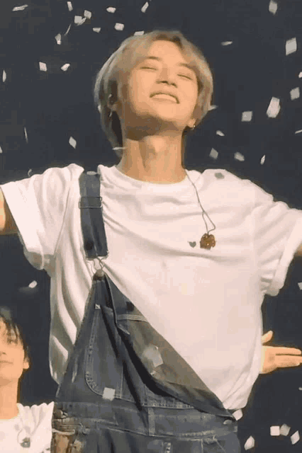 a man wearing overalls and a white t-shirt is standing in front of a crowd with his arms outstretched .