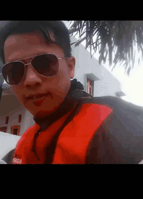 a man wearing sunglasses and a red jacket is standing in front of a house .