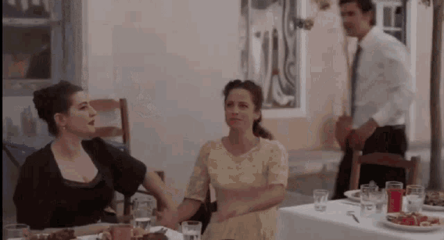 two women are sitting at a table with a man in a suit standing behind them .
