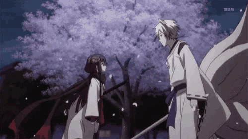 a man and a woman are standing in front of a cherry blossom tree with mbs on the bottom right