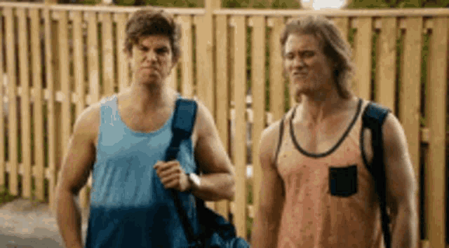 two men wearing tank tops and backpacks are standing next to each other in front of a wooden fence .