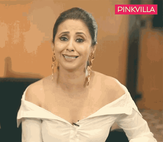 a woman wearing a white shirt and gold earrings with a pinkvilla logo behind her