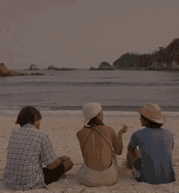 three people sit on a beach looking at the ocean